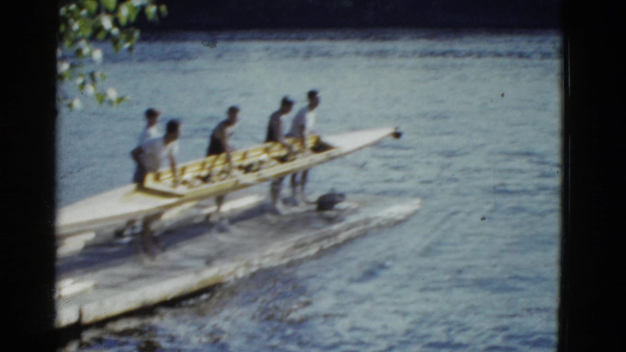Young men in swimwear and t-shirts move a long rowing shell along a waterfront dock in the supplied 1975 Boston footage.