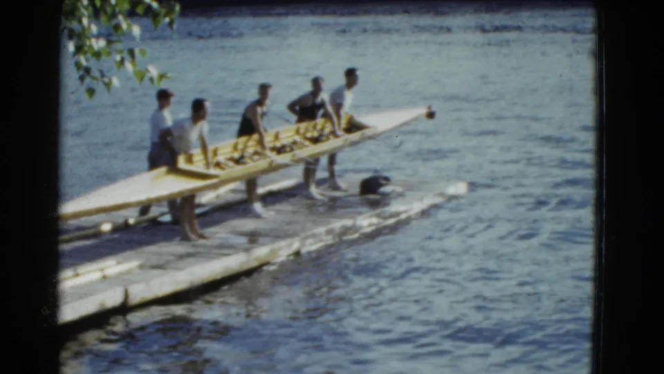 The 8mm frame shows young men gathered beside a long rowing shell on a dock, with water and overhanging leaves behind them.