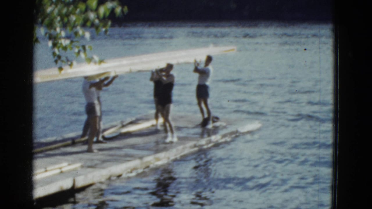 1975 Boston-area home-movie frame with several people holding a long rowing shell on a lakeside dock.