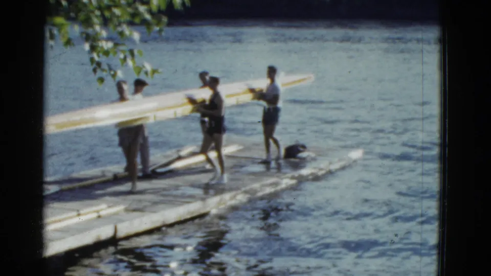 Several people in casual summer clothing carry a long rowing shell on a dock in 1975 Boston-area 8mm footage.