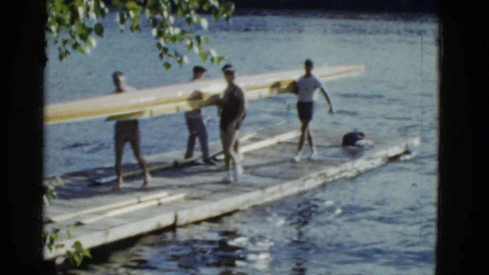 A 1975 Boston-area dock scene shows people walking with and standing near a long rowing shell beside the lake.