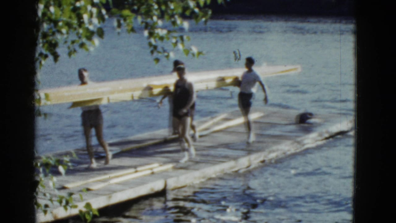Rowers stand on a wooden dock beside a yellow shell at the water's edge.