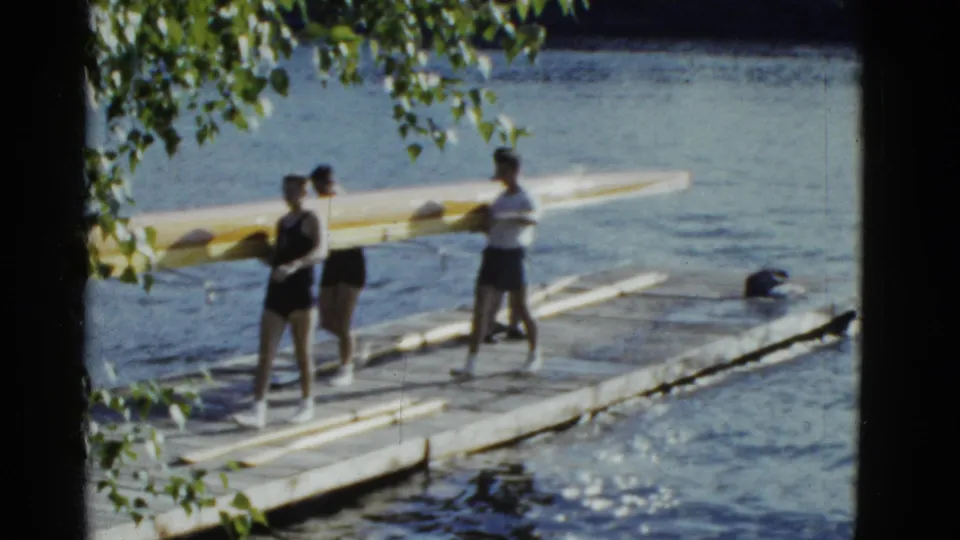 Rowers in athletic clothing carry a yellow racing shell along a wooden dock in the 1975 Boston clip.