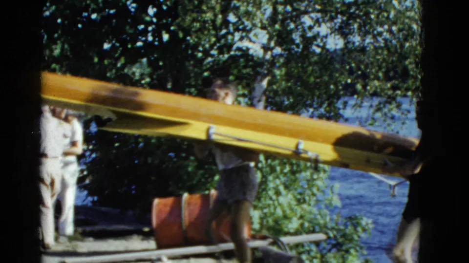 The yellow rowing shell fills the foreground as rowers carry it past leafy shoreline and dockside gear.