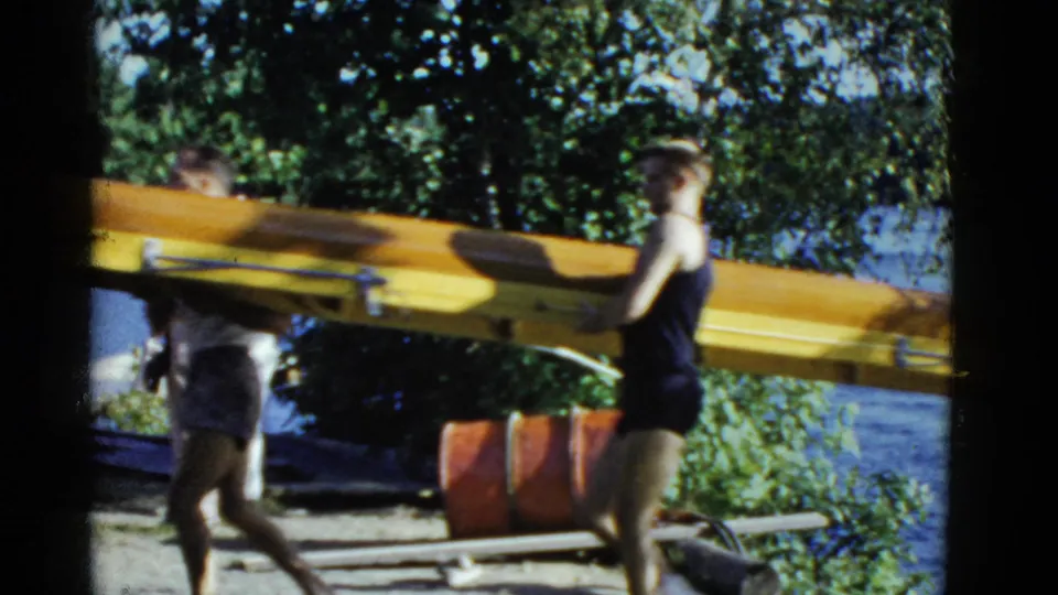 A rower carries the stern area of a yellow shell near the waterfront, with trees and gear visible behind.