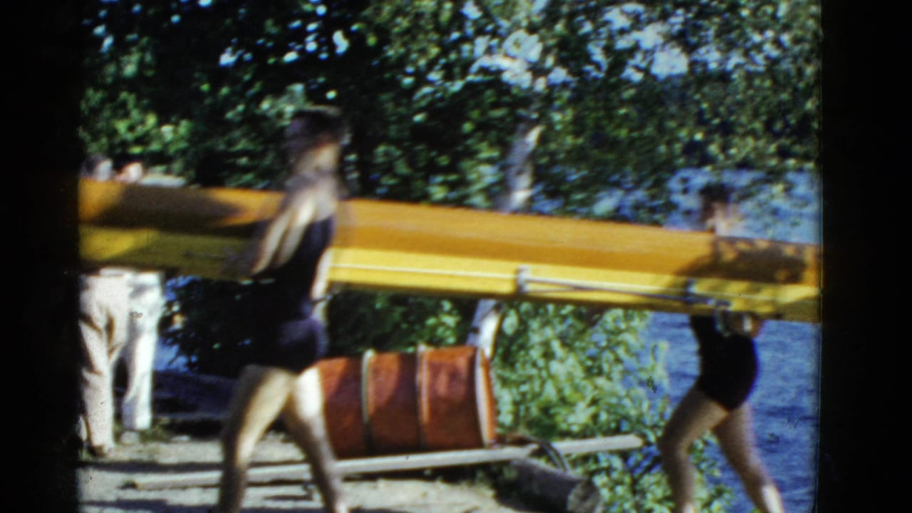 People carry a long wooden rowboat along a shaded lakeside path.