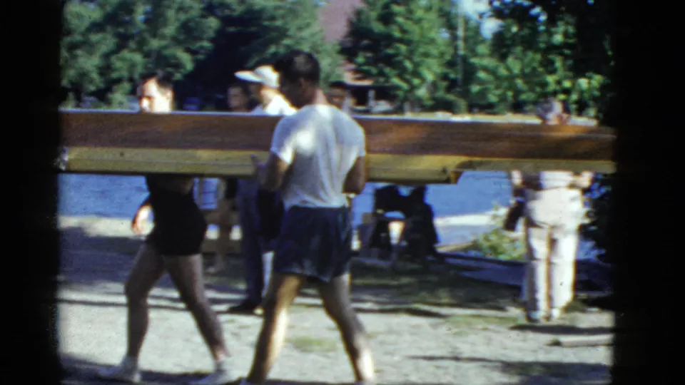 Several people in 1970s swimwear carry a long wooden rowboat along a Boston lakeside path with trees behind them.