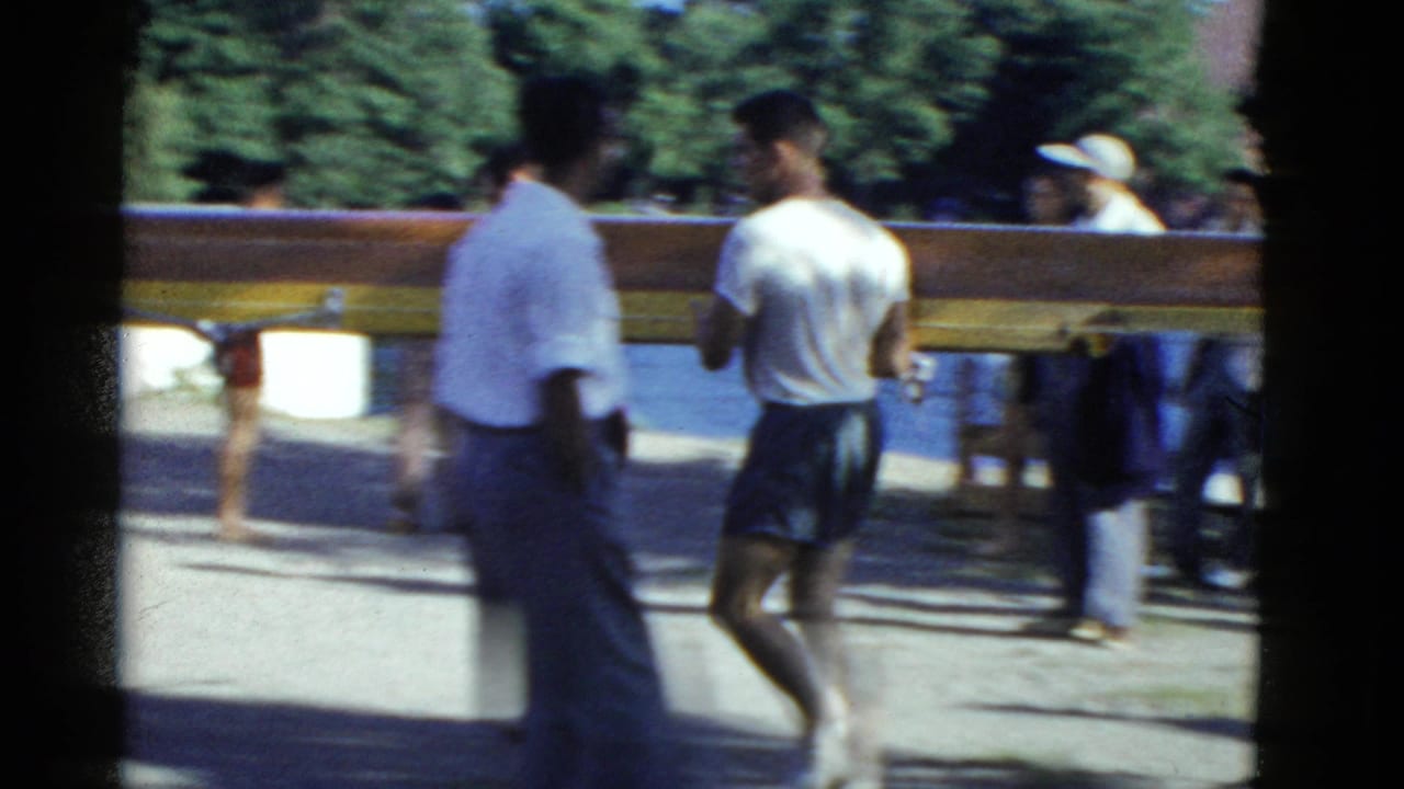 1975 Boston-area home-movie frame showing people beside a long rowing shell near trees and water.