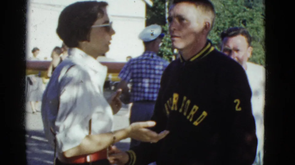 1975 Boston-area home-movie frame of men conversing near a rower group and a long shell in bright sunlight.