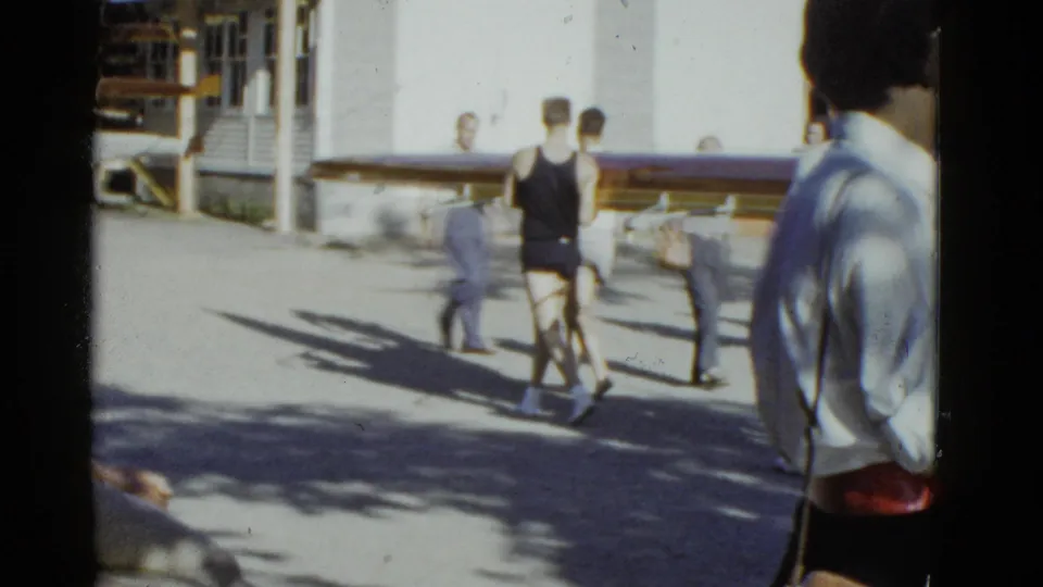 1975 Boston-area frame showing rowers carrying a long shell along a sunlit paved path near a building and onlookers.