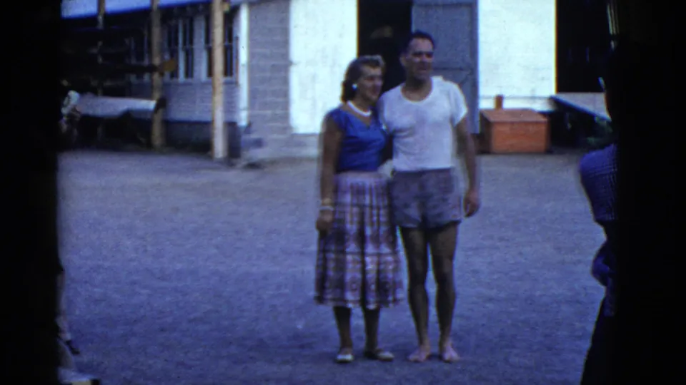 1975 Boston home-movie still of a couple standing side by side, with the woman in a blue top and the man in a white t-shirt and swim trunks.