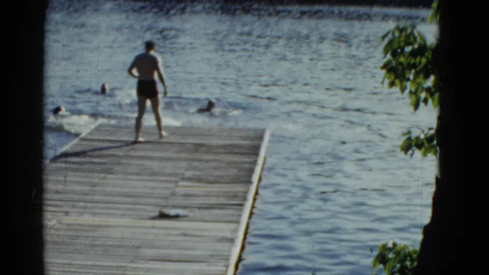 A man stands near the end of a wooden dock as swimmers gather in the lake, with tree leaves at the edge of the 8mm frame.