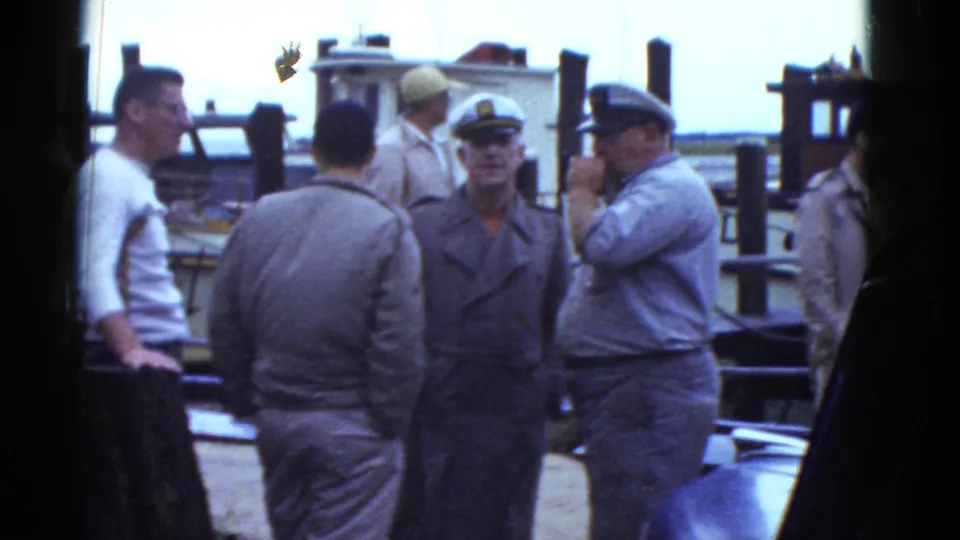 Several adult men stand close together on a Boston waterfront dock, with boats and pilings behind them.