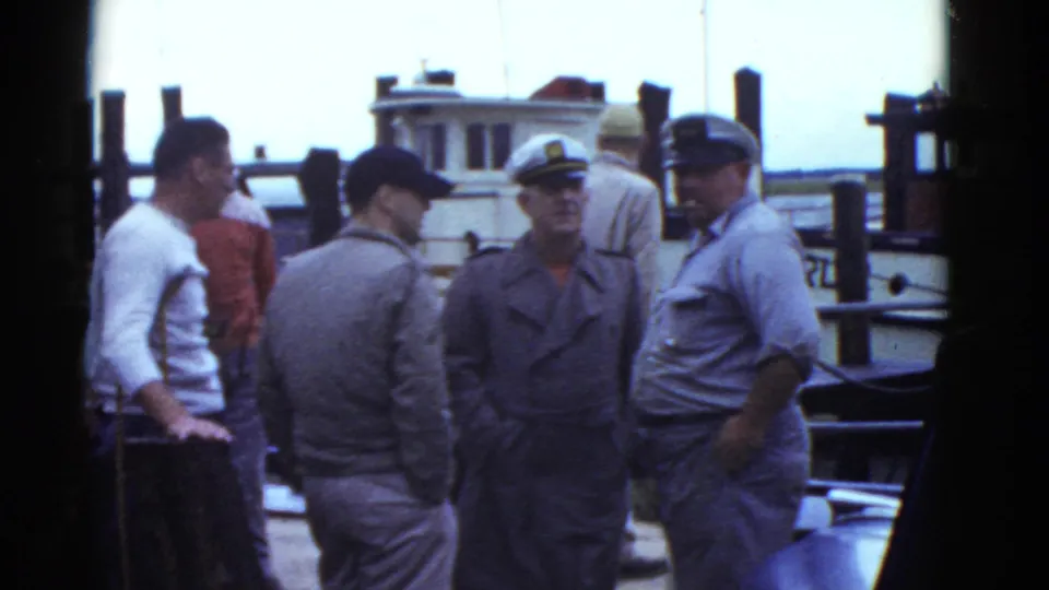 The group remains on the Boston dock in 1975, with one man facing away and others framed by boats and pilings.