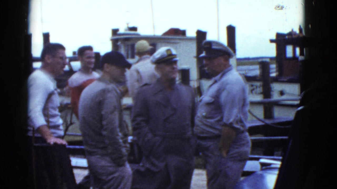 Men in uniforms and caps talk on a dock beside a small boat.