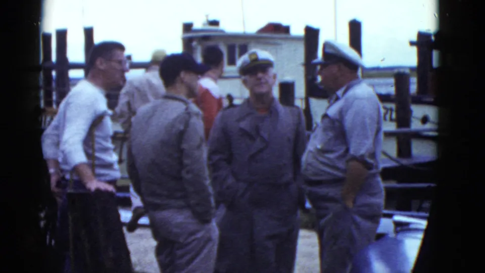 In Boston harbor footage from 1975, men in gray uniforms and caps converse near a small docked vessel.