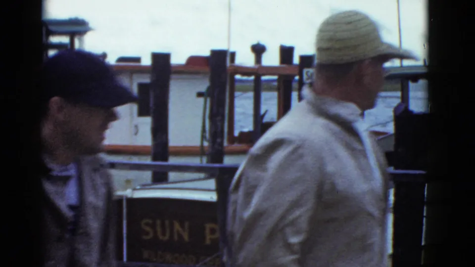 Two men pass close to the camera on a 1975 Boston dock, with moored boats, pilings, water, and signage in the background.