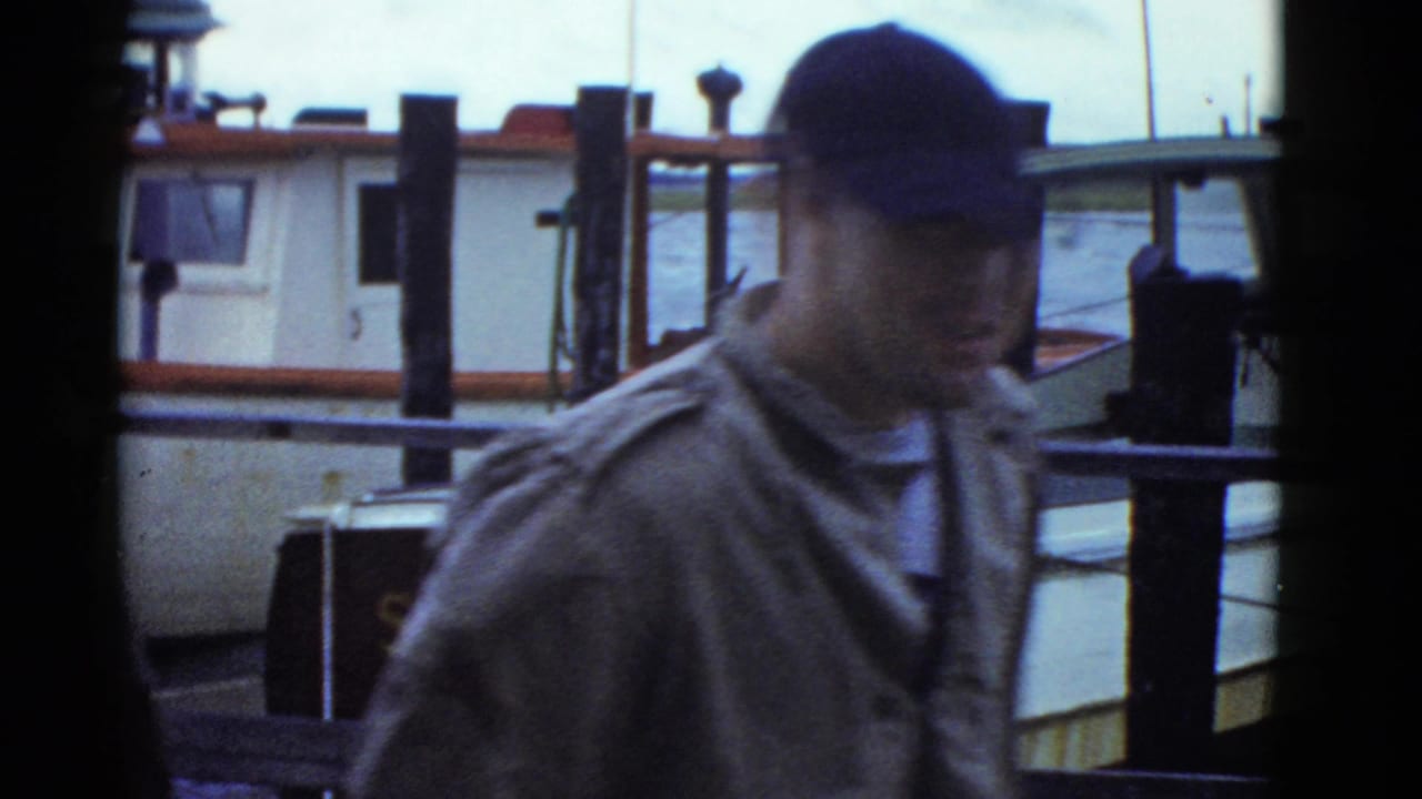 In supported 1975 Boston home-movie footage, a man in a dark cap moves past dock railings with cabin boats behind him.