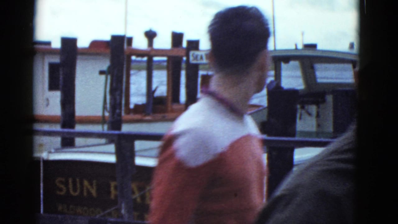 A man in a red and white shirt stands near Boston dock railings, with boat cabins and partial SEA WOLF signage behind him.