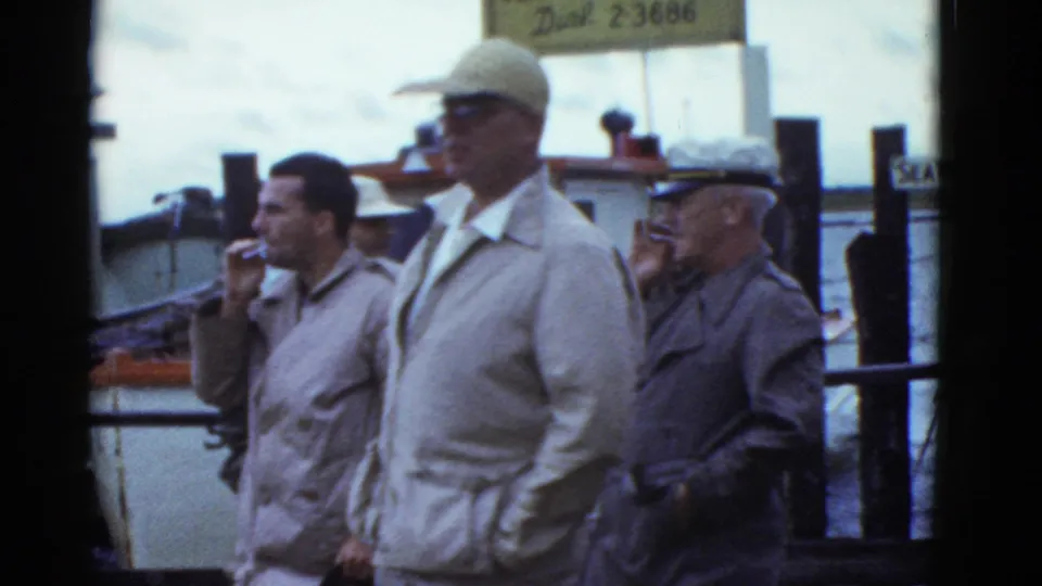 Three adult men stand by Boston dock railings in 1975 footage, one smoking, with moored boats and a sign above them.