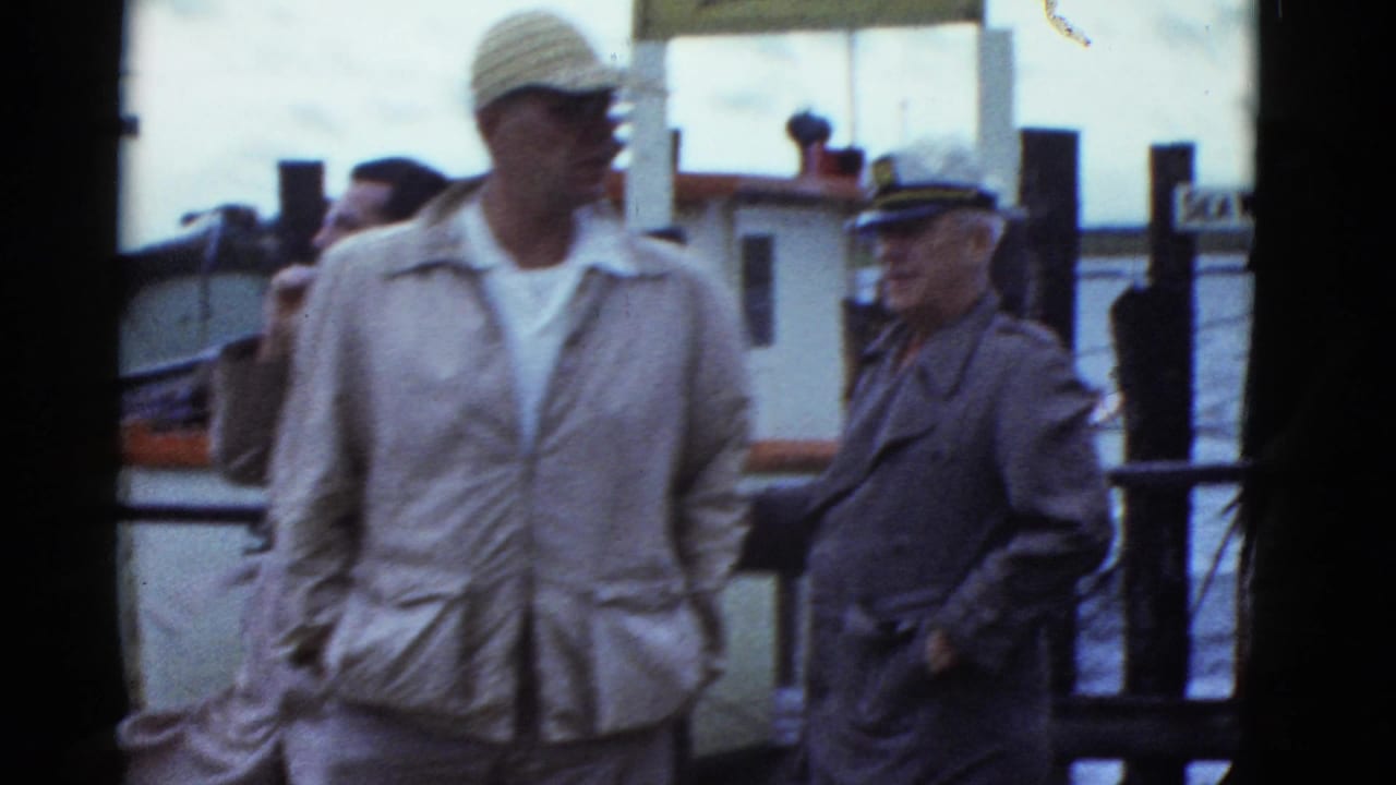 Men in light jackets and caps stand on a pier near water and a boat.