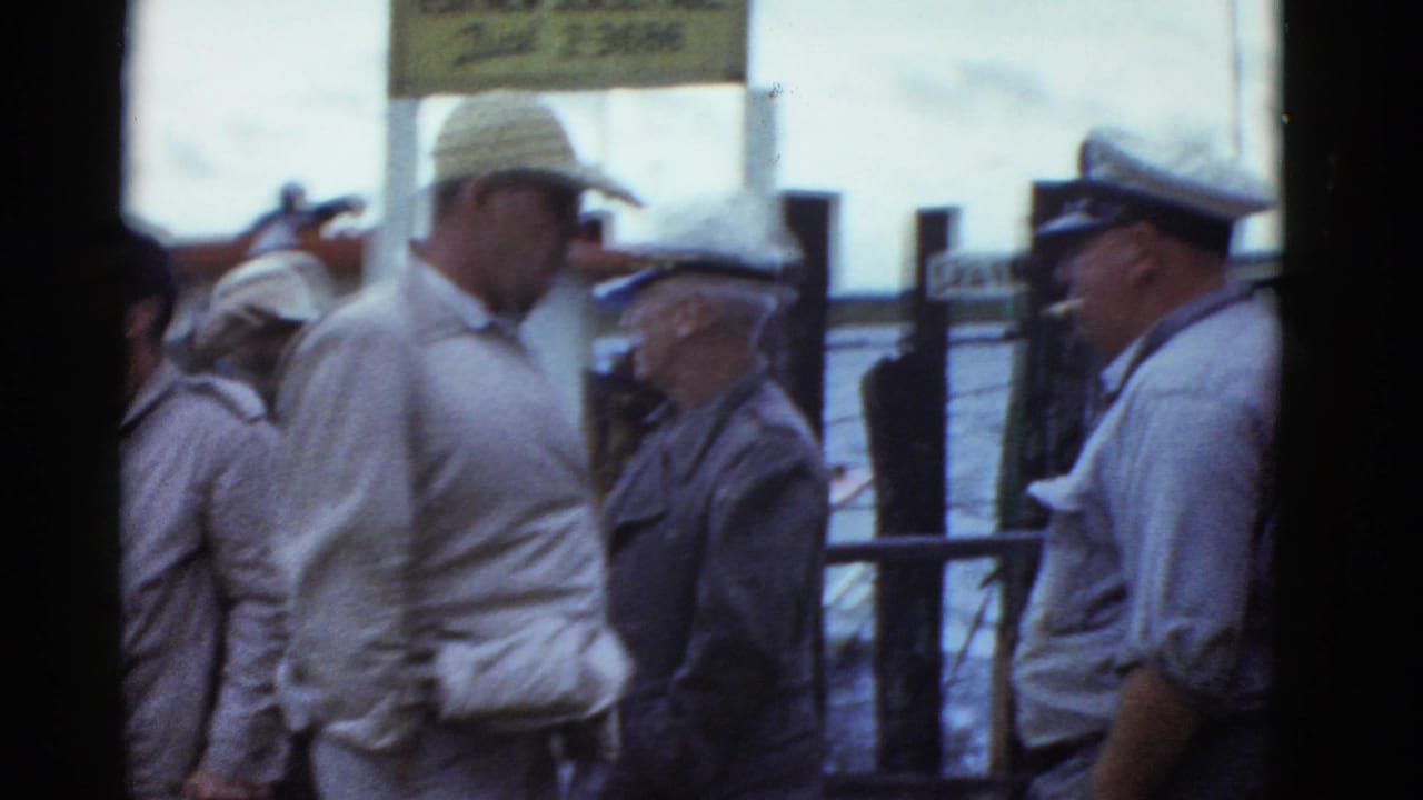 A man in a light jacket faces other men beside a waterfront railing.