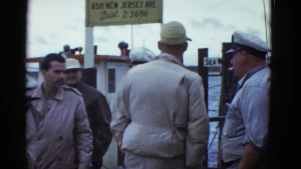 1975 Boston waterfront frame showing men in jackets near pier railings, water, and a partly legible address sign overhead.