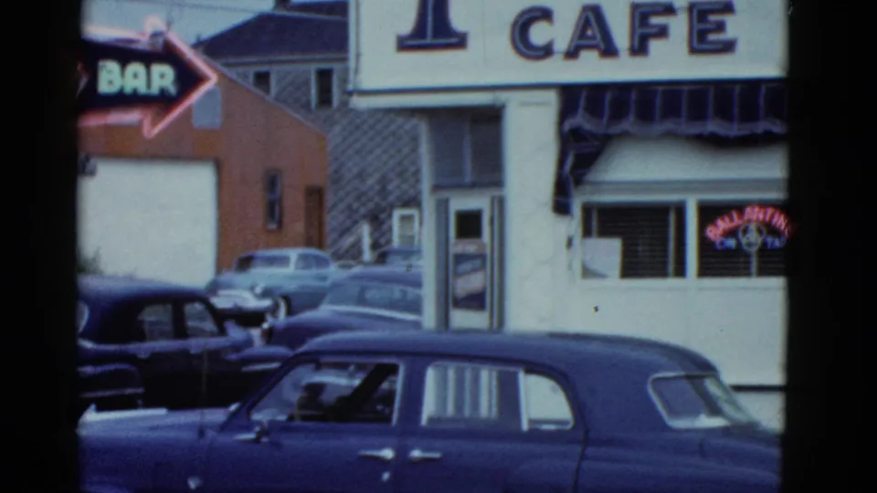 Boston 1975 frame with a red BAR arrow sign, partial cafe facade, striped awning, and a dark blue older sedan in front.