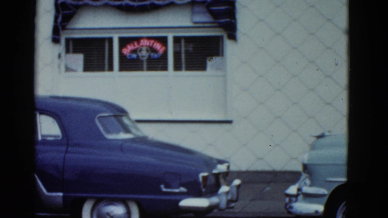 Blue sedan and pale turquoise car sit along a white storefront wall in 1975 Boston-area home-movie footage.