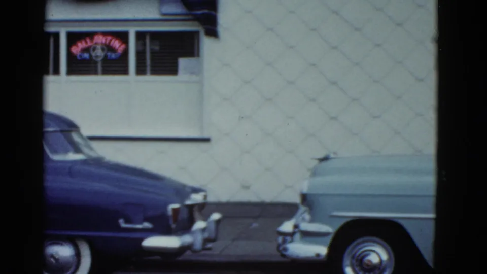 A passing 8mm view shows parts of two parked cars against a white storefront with a neon sign and textured wall.