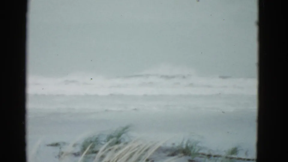 1975 8mm coastal frame showing a muted shoreline, white wave break, and dune grass along the lower foreground.