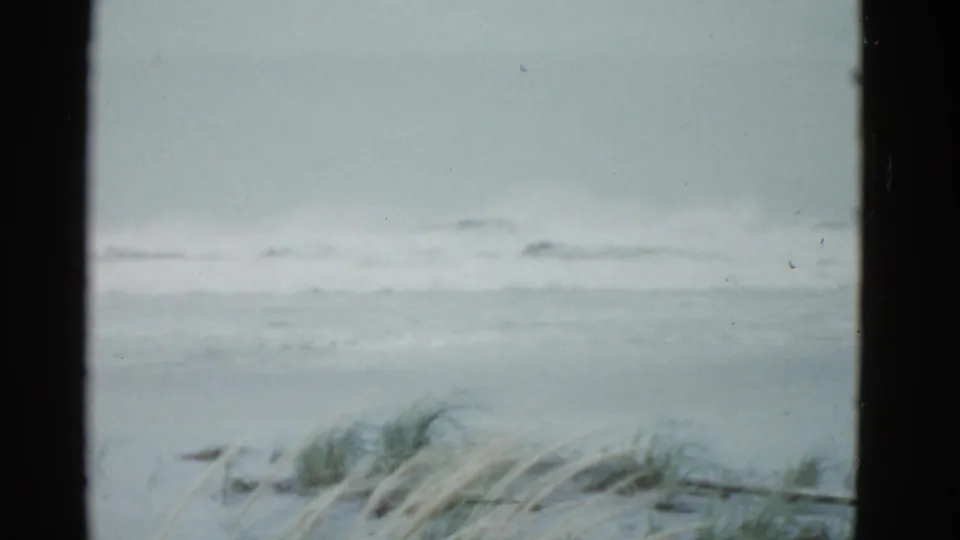 Boston 1975 beach frame with calm ocean tides, sandy foreground, and wind-bent grass beneath a gray sky.