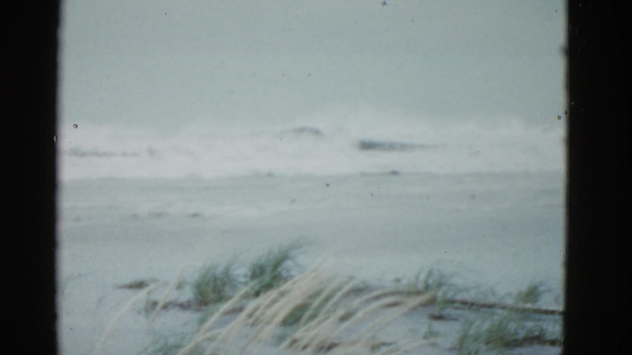 1975 Boston-area 8mm frame showing gray surf, white wave foam, beach grass, and dark film-frame borders.