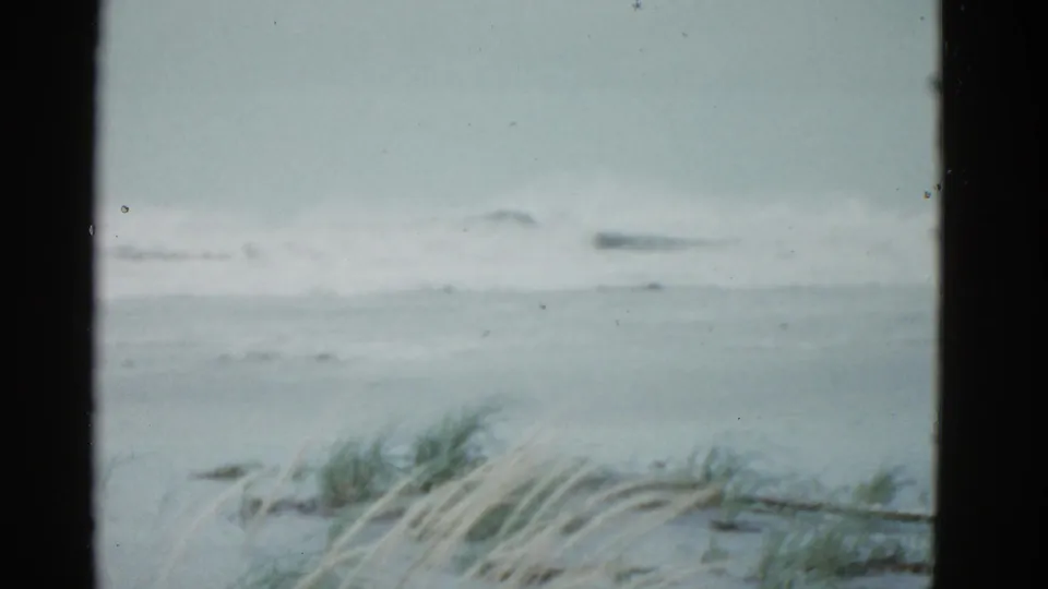 1975 Boston-area 8mm frame showing gray surf, white wave foam, beach grass, and dark film-frame borders.