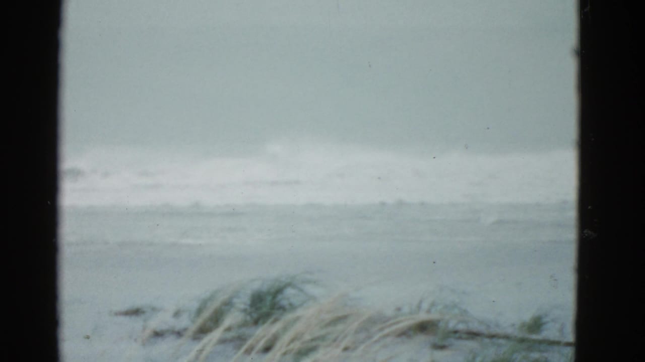 1975 Boston-area coastal frame with muted gray waves, windblown grass, overcast sky, and visible 8mm frame borders.