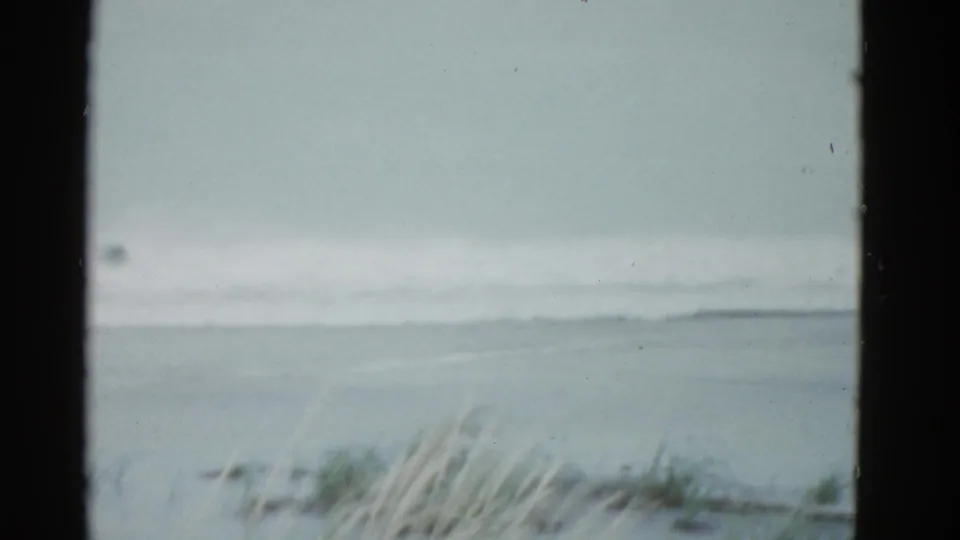 Cautious Boston-area 1975 coastal view on 8mm film, showing rough water, a gray horizon, and foreground dune grass.