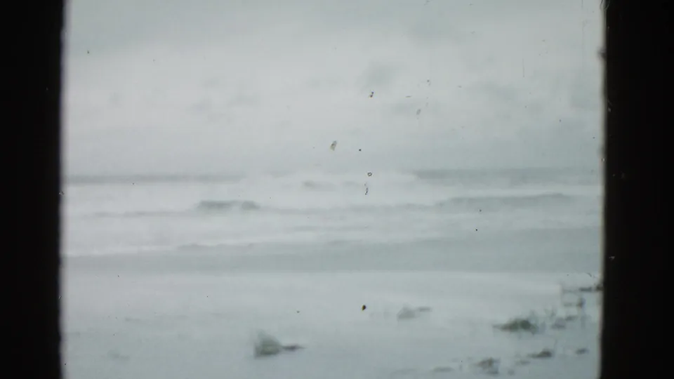 1975 8mm coastal home-movie frame showing rough waves under a gray sky, with sparse beach grass and no visible people.