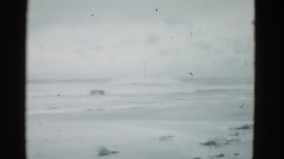 1975 8mm home-movie view of an empty Boston-area beach with pale waves, cloudy sky, and dark foreground framing.