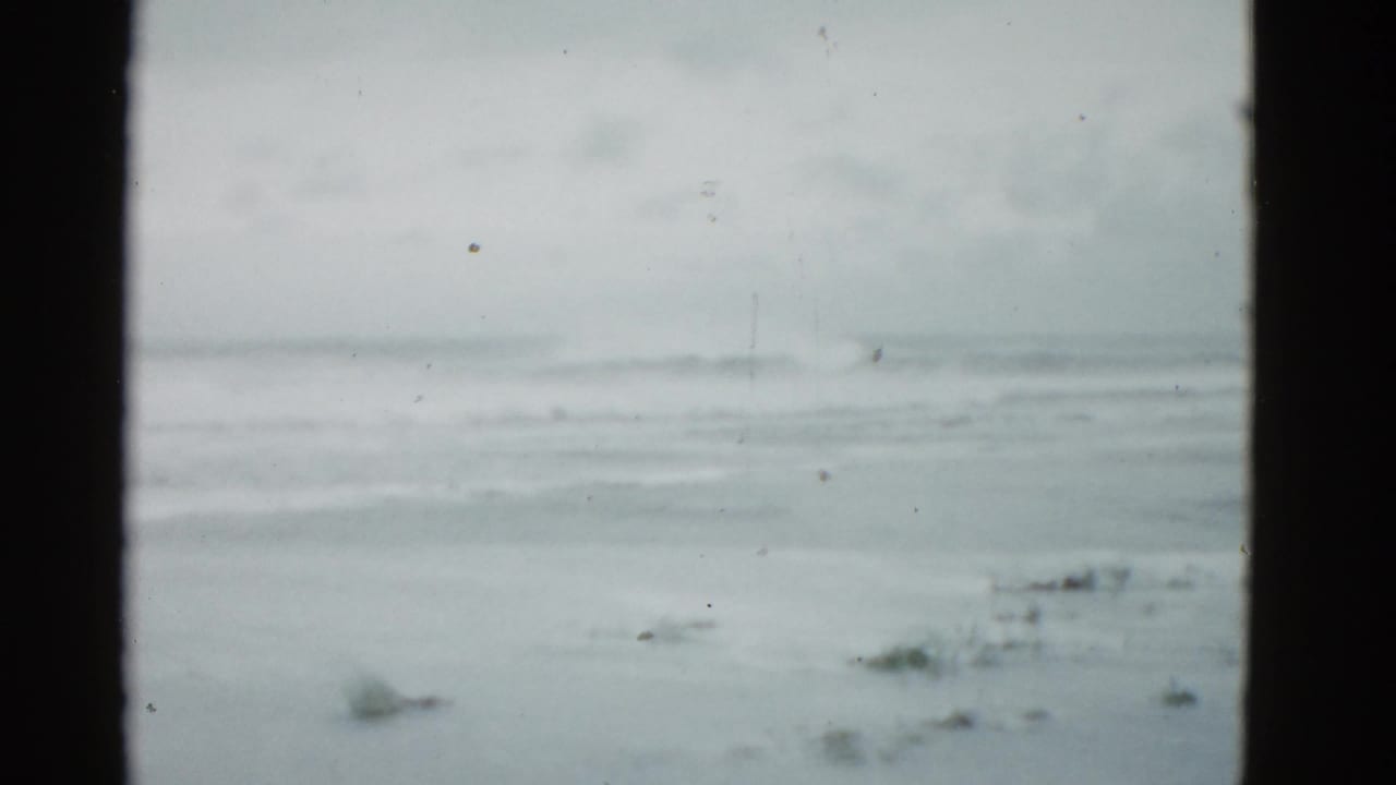 Overcast 1975 8mm coastal view near Boston, with low surf, wet sand, and no visible people or structures.