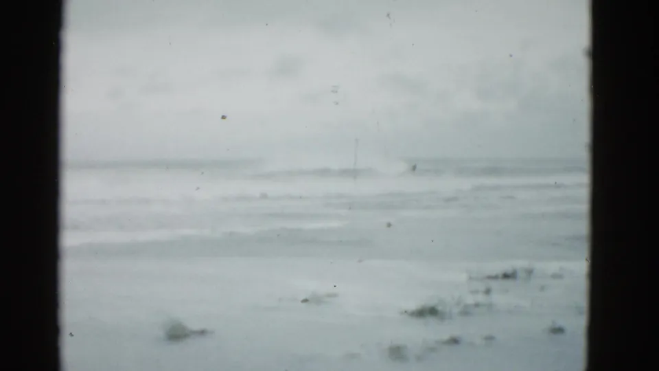 Overcast 1975 8mm coastal view near Boston, with low surf, wet sand, and no visible people or structures.