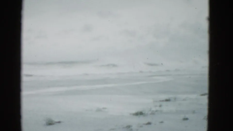 1975 home-movie shoreline frame near Boston, with distant waves, empty sand, cloudy light, and visible film gate edges.