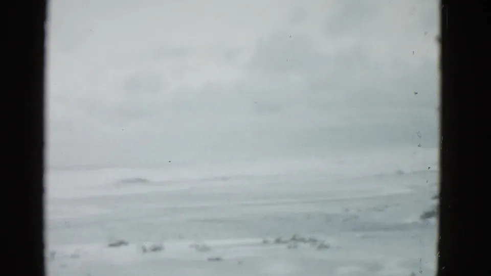 1975 Boston-area 8mm still showing a frozen lake under a washed winter sky, framed by dark gate edges and visible dust.