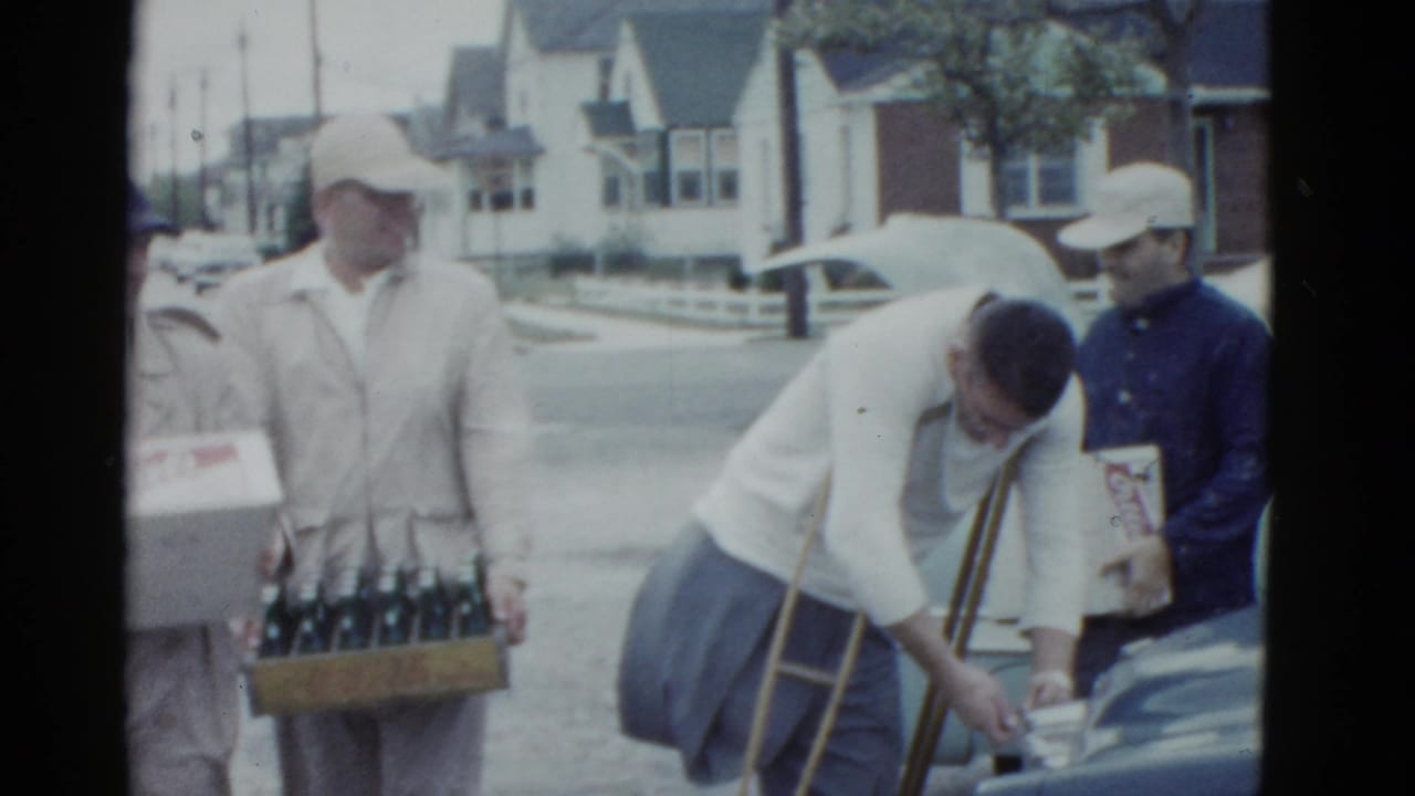 In 1975 Boston home-movie footage, adult men stand near a parked car while a man with crutches bends toward items beside the vehicle.