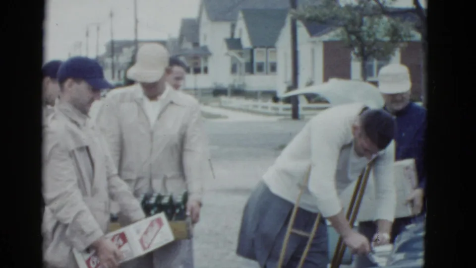 A 1975 Boston street frame shows men with boxes and bottle crates around a parked car, including one man using crutches as he reaches down.
