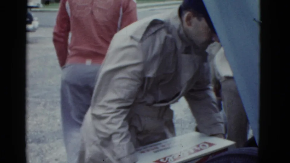 A man in a light jacket and dark cap handles an Ortlieb's beer box beside a car during the Boston 1975 home-movie clip.