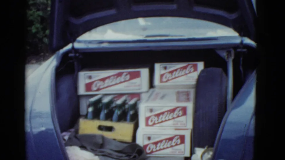1975 Boston-area 8mm frame showing a blue car trunk filled with Ortlieb beer boxes, a wooden bottle crate, fabric, and a spare tire.