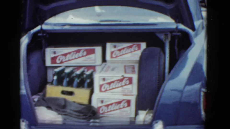 Soft 8mm frame of the blue car trunk, with stacked red-and-white beer boxes, a crate of bottles, and a spare tire visible at right.