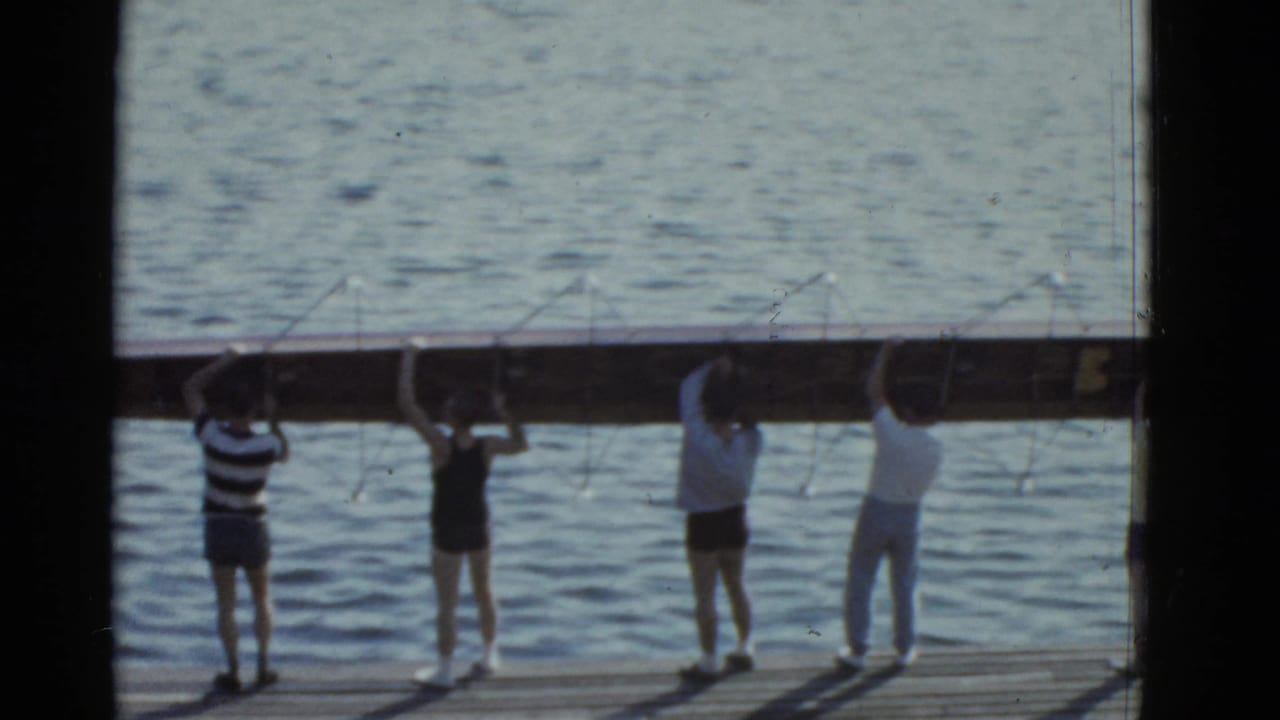 Men stand on a dock holding a long wooden log beside lake water.