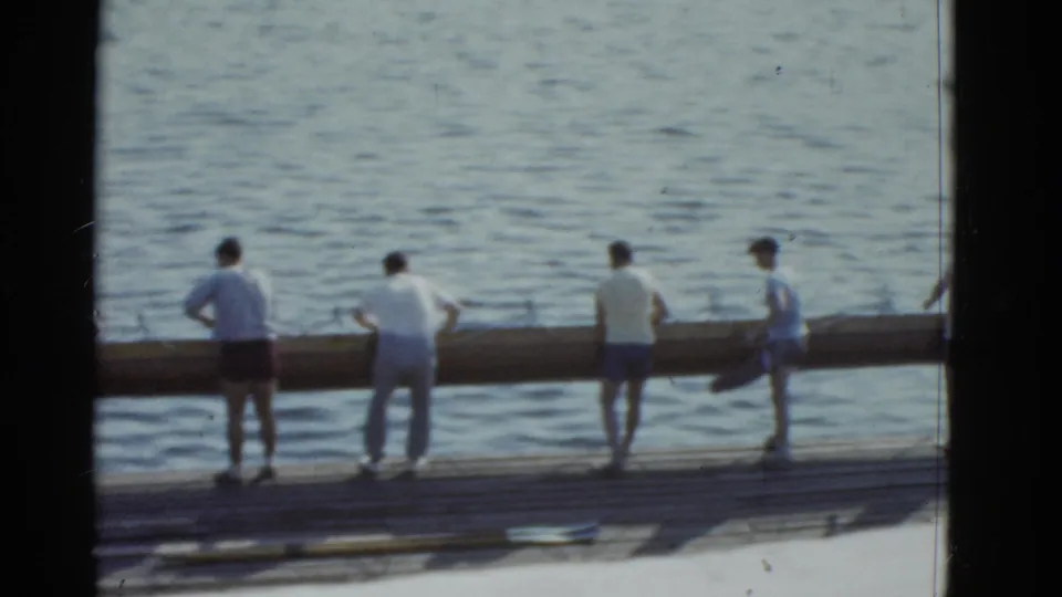 1975 Boston-area home-movie frame showing men spaced along a dock as a long log rests near the lake water.