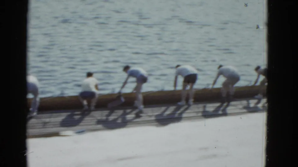 1975 Boston-area 8mm frame showing several men bending over a long log on a dock, apparently adjusting or preparing it near the water.